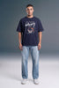 A man wearing a navy blue t-shirt with a white and red basketball graphic that reads 'AUTHENTIC CHAMPION', paired with light blue jeans and white sneakers, standing against a gray studio background.
