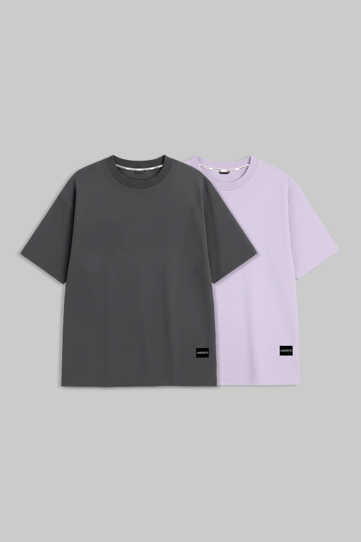 Two short-sleeve crewneck t-shirts displayed side-by-side on a solid gray background. The front t-shirt is dark charcoal gray, and the t-shirt behind it is light lavender purple.