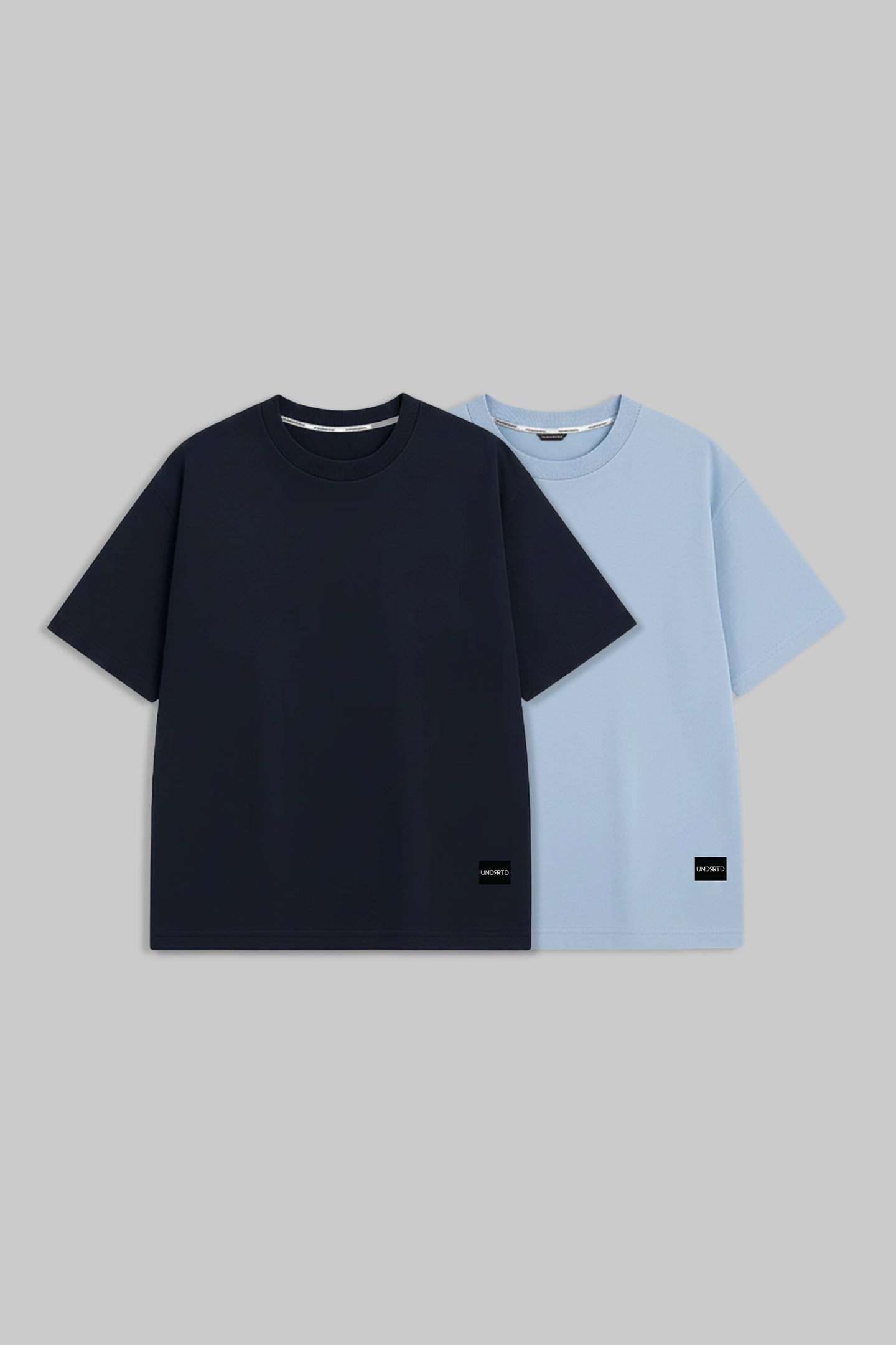 Two short-sleeve crew-neck t-shirts displayed flat against a solid gray background. The front t-shirt is solid navy blue, and the t-shirt behind it is solid light blue.