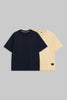 Two short-sleeve crew-neck t-shirts displayed flat against a solid gray background. The front t-shirt is dark navy blue, and the t-shirt behind it is a light cream/beige color.