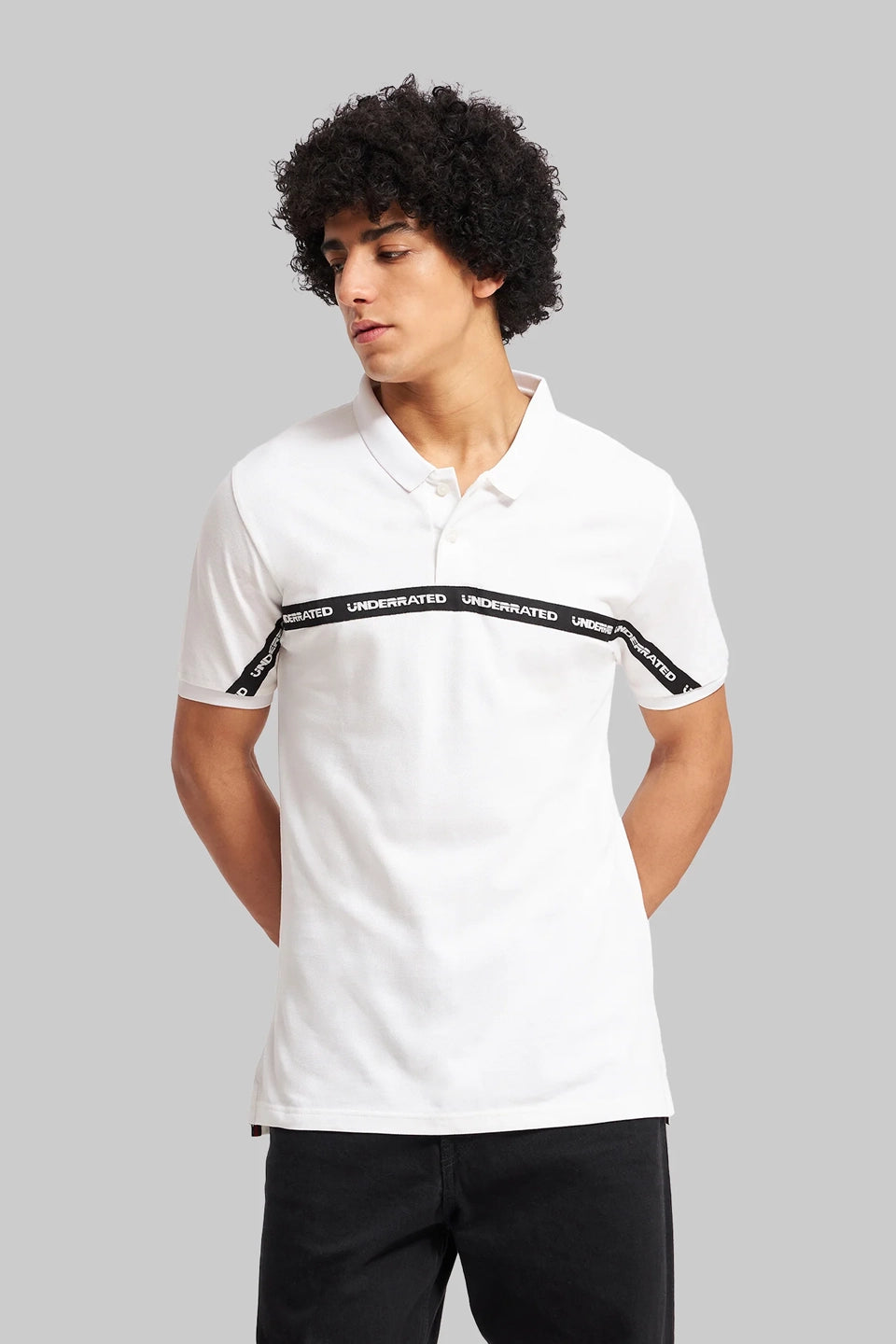 A man with curly hair wearing a white polo shirt featuring a black graphic "UNDERRATED" band across the chest.