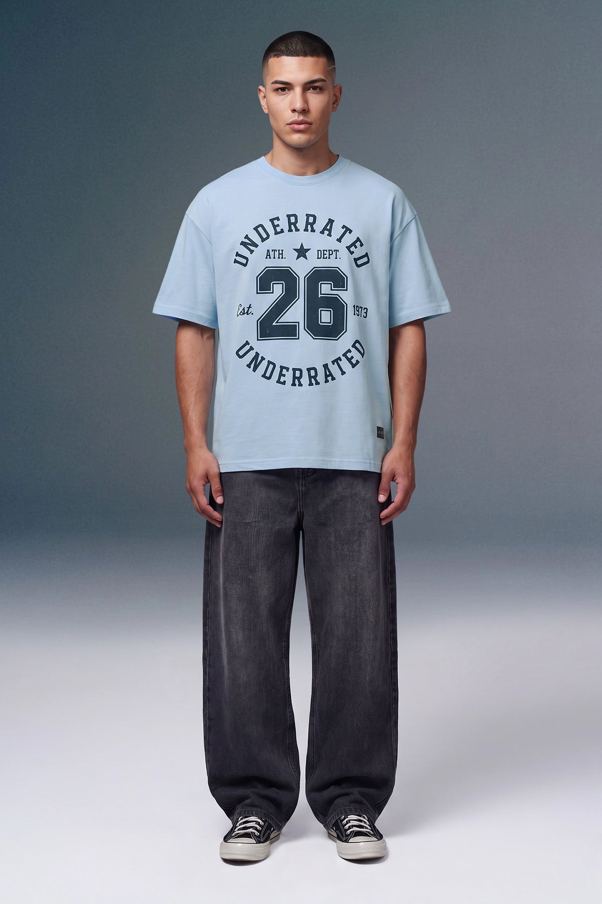 A male model stands against a gray studio background wearing a light blue graphic T-shirt with dark navy text reading 'UNDERRATED 26 UNDERRATED' and dark gray baggy jeans with black sneakers.