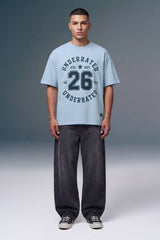 A male model stands against a gray studio background wearing a light blue graphic T-shirt with dark navy text reading 'UNDERRATED 26 UNDERRATED' and dark gray baggy jeans with black sneakers.