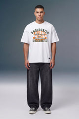 A male model stands against a gray studio background wearing a white graphic t-shirt with orange and black text, dark gray baggy jeans, and black sneakers.