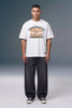 A male model stands against a gray studio background wearing a white graphic t-shirt with orange and black text, dark gray baggy jeans, and black sneakers.