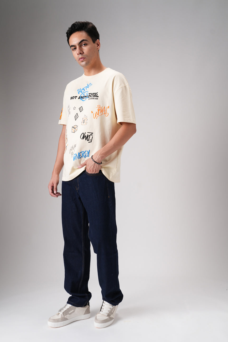 Full-body view of man in beige graphic t‑shirt and dark jeans standing straight in studio with neutral backdrop.
