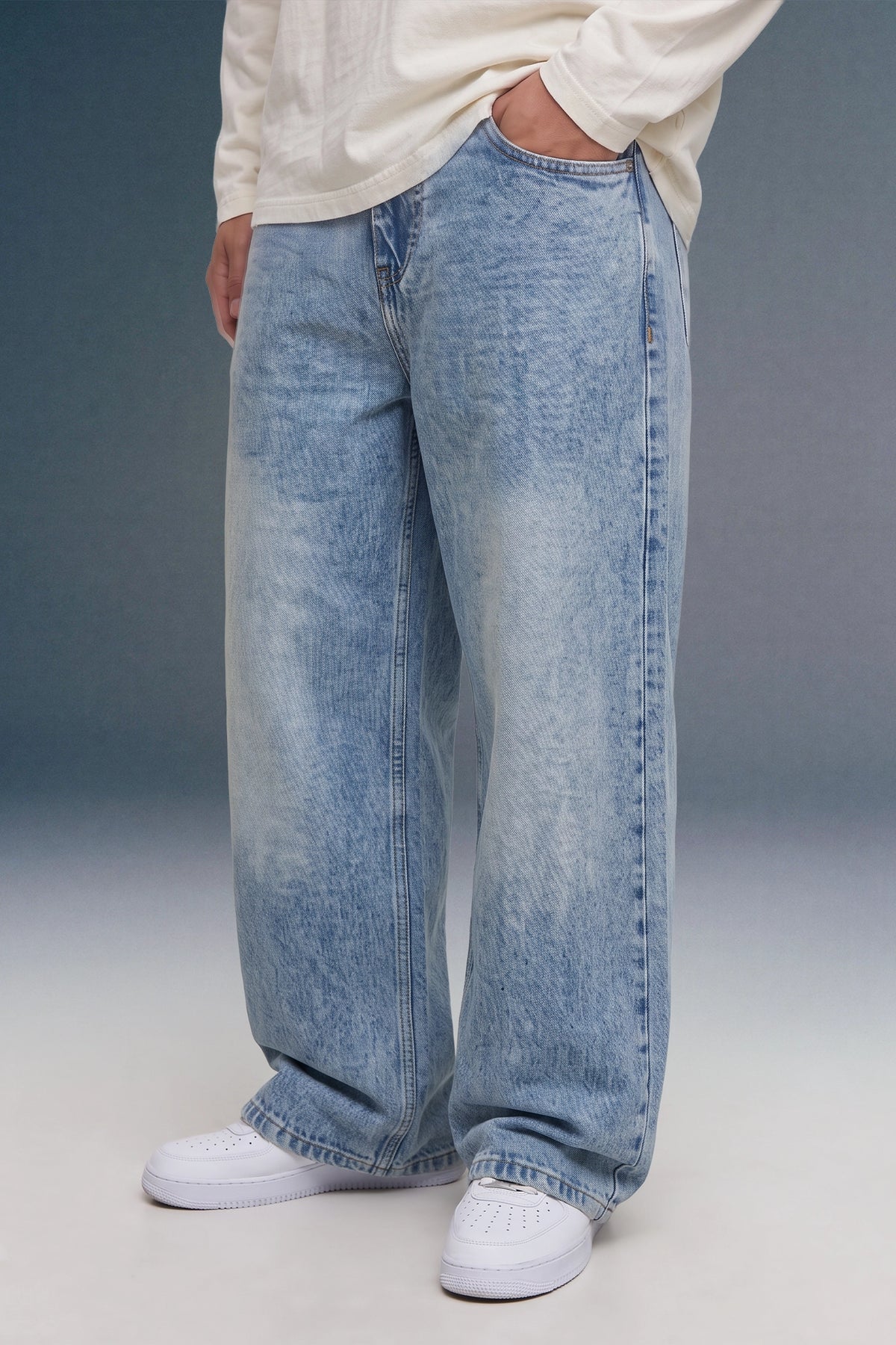 A person wearing light-wash, wide-leg denim jeans with a faded wash, styled with a cream long-sleeve top and white sneakers against a studio backdrop.