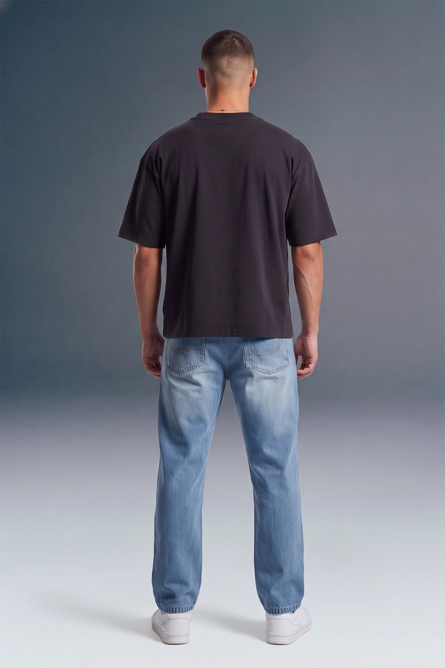 A man viewed from behind wearing a dark charcoal gray t-shirt, light blue jeans, and white sneakers, standing against a dark gray studio background.