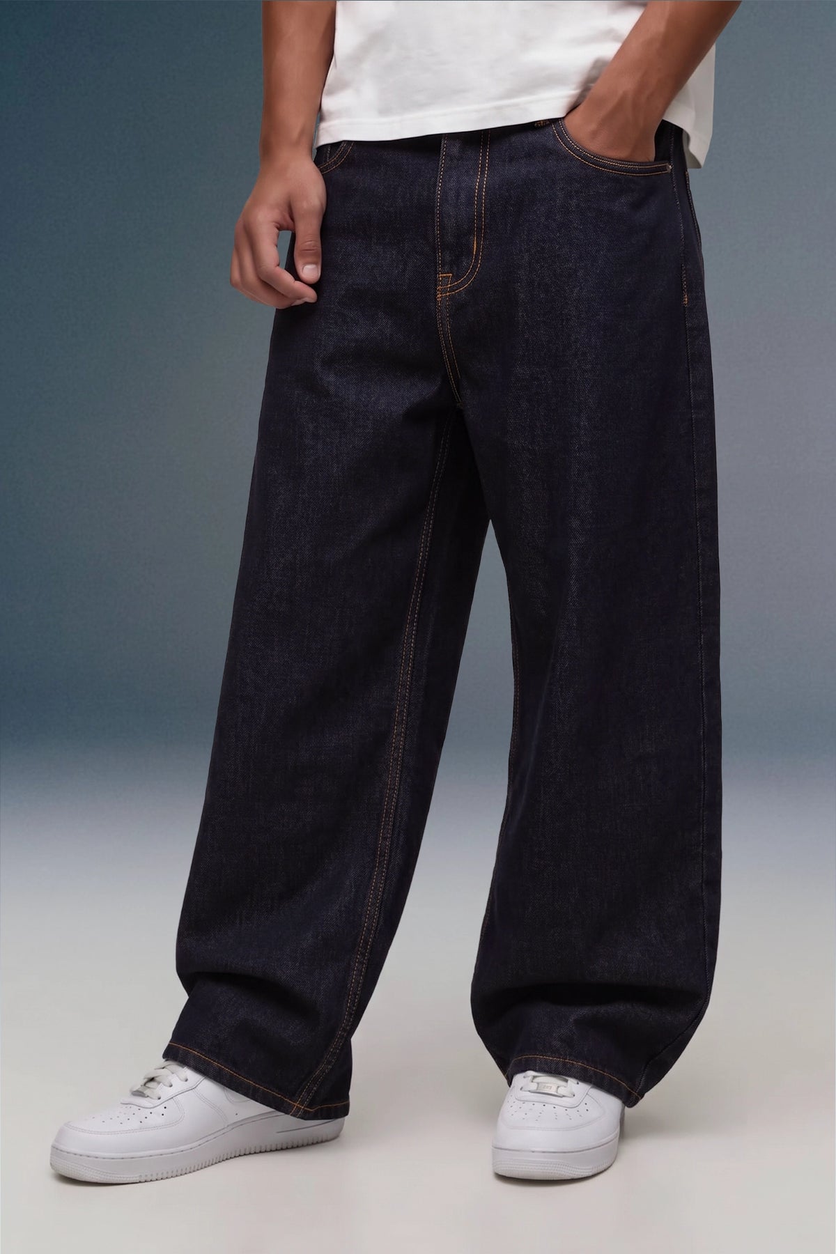 A person wearing very wide-leg dark indigo denim jeans with contrast tan stitching, styled with a white t-shirt and white sneakers against a studio backdrop.