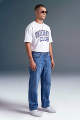 A man wearing a white graphic t-shirt with blue lettering that reads 'UNDERGRADUE CLUB', paired with blue jeans and white sneakers, standing against a gray studio background.