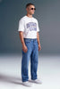 A man wearing a white graphic t-shirt with blue lettering that reads 'UNDERGRADUE CLUB', paired with blue jeans and white sneakers, standing against a gray studio background.