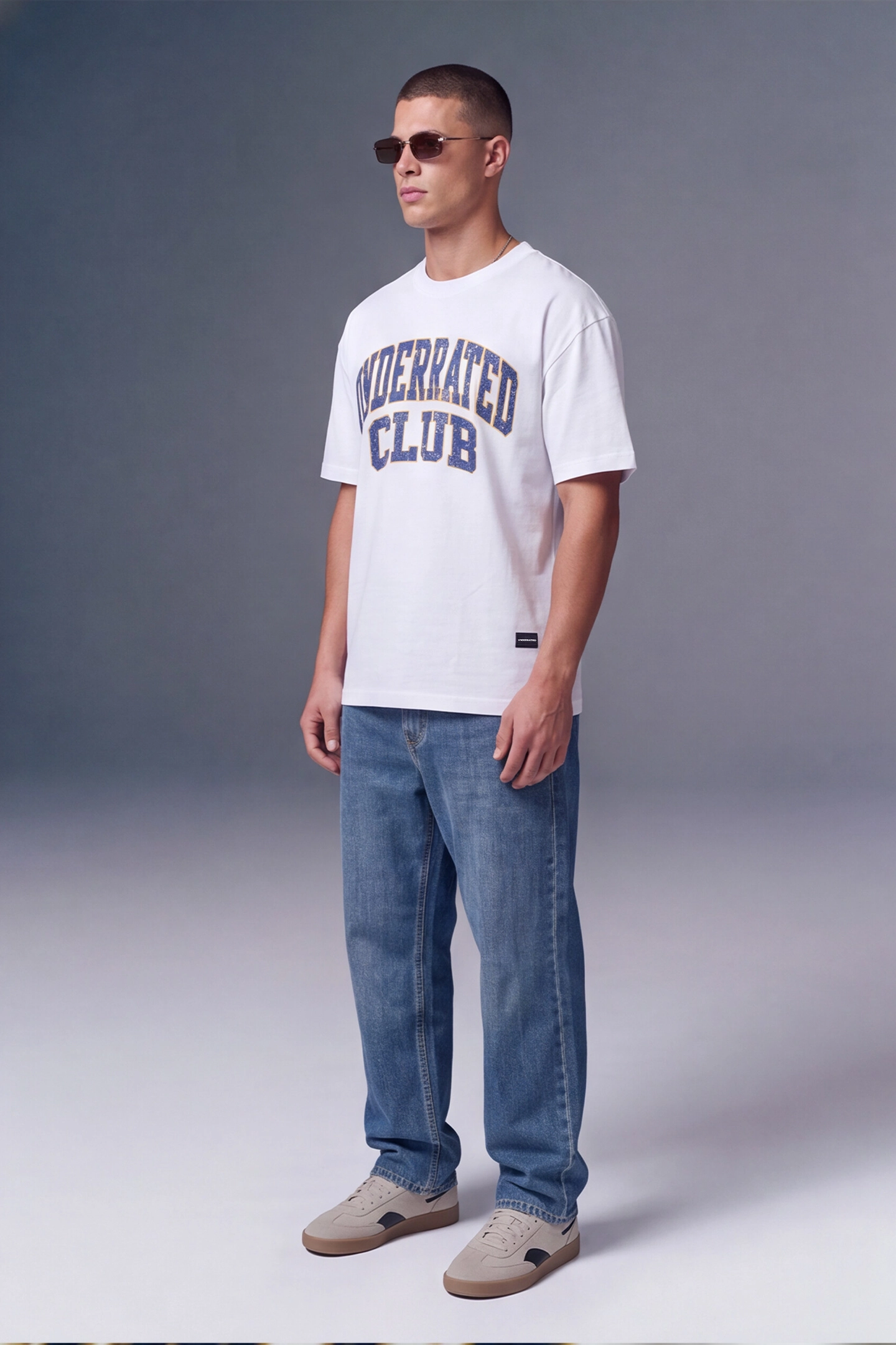 A man wearing a white crewneck t-shirt with 'MODERATED CLUB' printed in navy blue arched text on the chest, paired with blue jeans and beige sneakers, standing against a gray studio background.