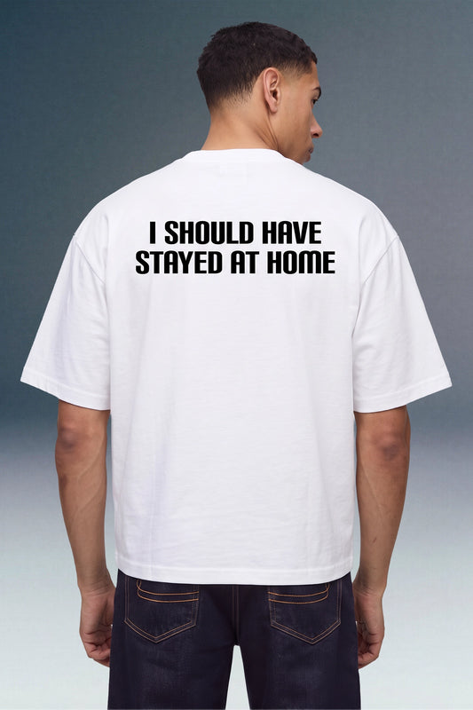A man viewed from behind wearing a white t-shirt with the text 'I SHOULD HAVE STAYED AT HOME' printed in bold black capital letters on the back, paired with dark blue jeans.