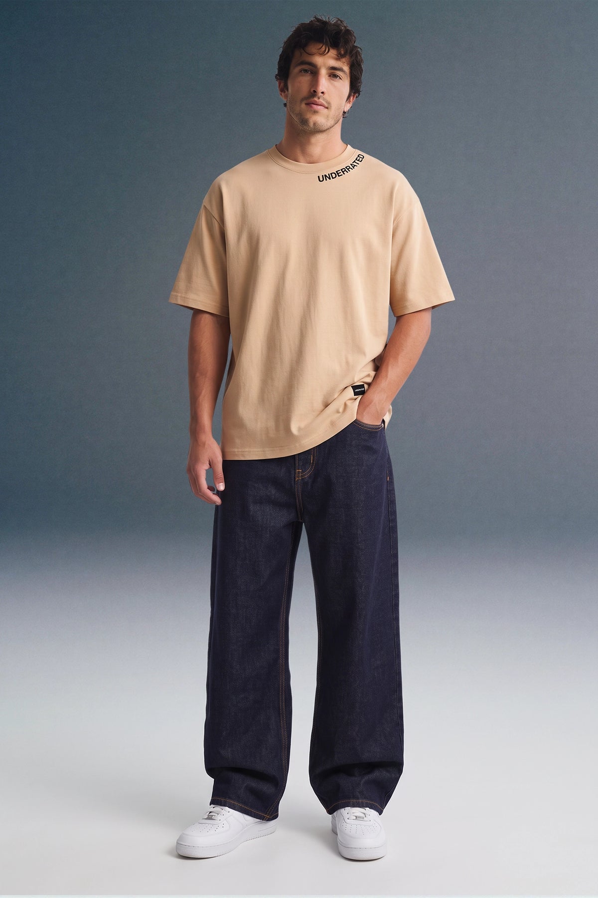Full-length front view of a man looking forward with a hand in his pocket, wearing a beige crewneck T-shirt with collar print detailing, dark navy pants, and white sneakers.