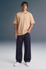Full-length front view of a man looking forward with a hand in his pocket, wearing a beige crewneck T-shirt with collar print detailing, dark navy pants, and white sneakers.