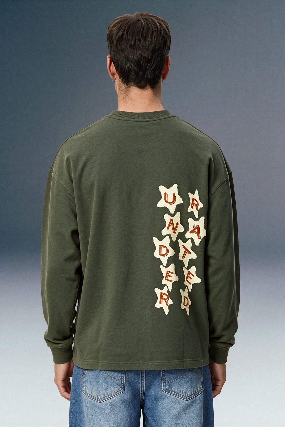 A man viewed from behind wearing an olive green long-sleeve shirt with a vertical graphic on the back made of cream star shapes containing the word 'UNLIMITED' in red letters, paired with blue jeans against a dark gray studio background.