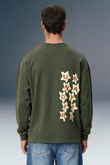 A man viewed from behind wearing an olive green long-sleeve shirt with a vertical graphic on the back made of cream star shapes containing the word 'UNLIMITED' in red letters, paired with blue jeans against a dark gray studio background.