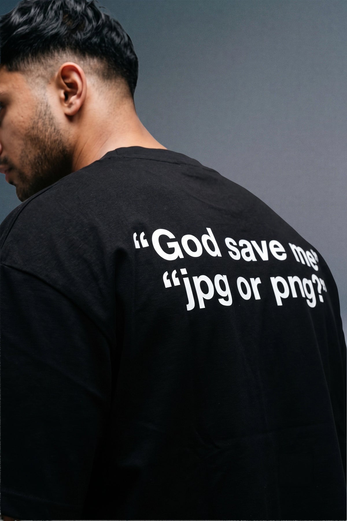 A man shown from the back wearing a black t-shirt with white text on the back that reads “God save me "jpg or png?” against a dark gray background.