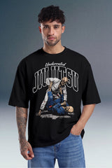 A man wearing a black graphic t-shirt with a white and blue 'Underrated Jiu Jitsu' print, paired with blue jeans, set against a dark gray studio background.