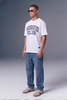 A man wearing a white crewneck t-shirt with 'MODERATED CLUB' printed in navy blue arched text on the chest, paired with blue jeans and beige sneakers, standing against a gray studio background.