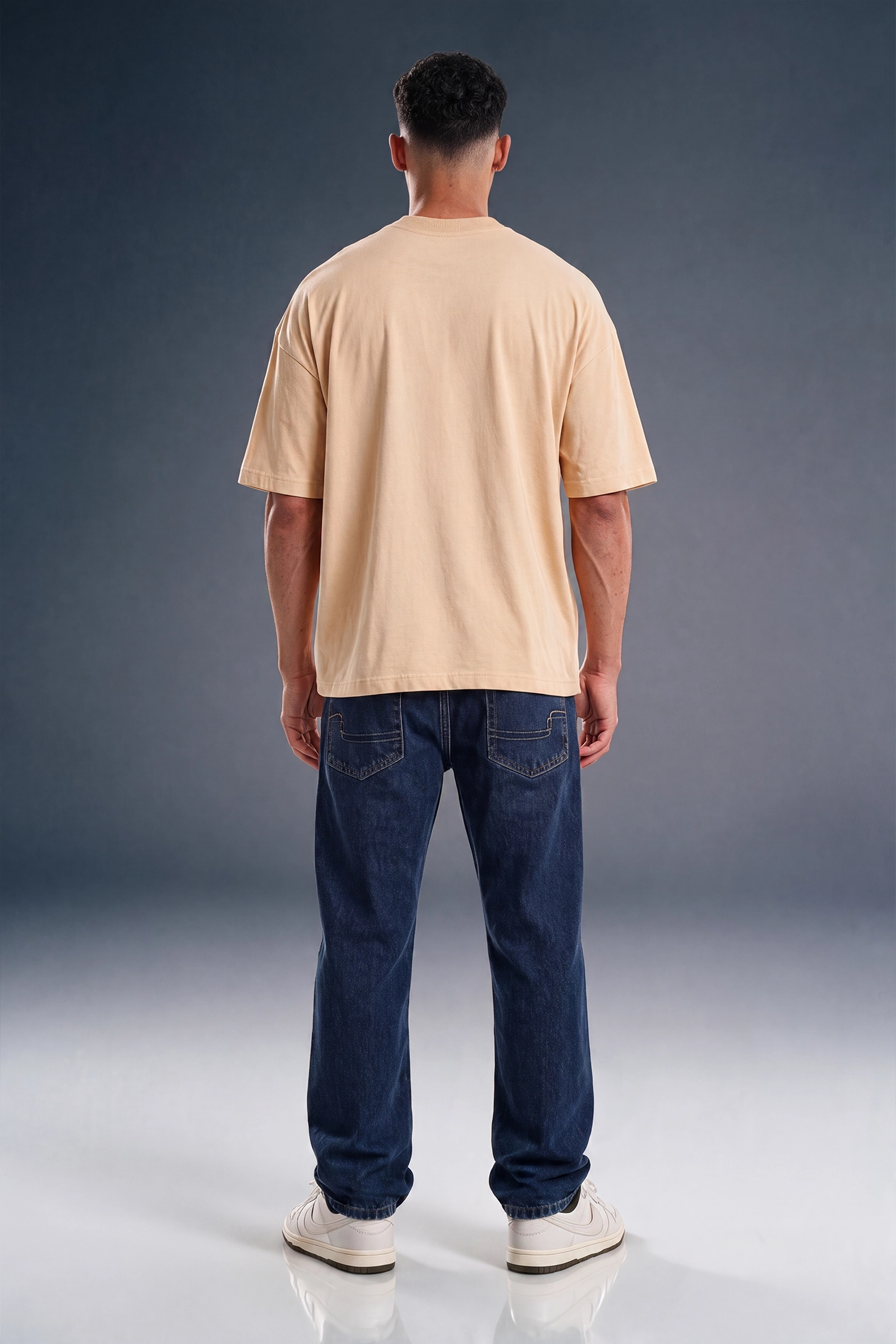 A man viewed from behind wearing a light beige oversized t-shirt, dark blue jeans, and white sneakers, standing against a dark gray studio background.
