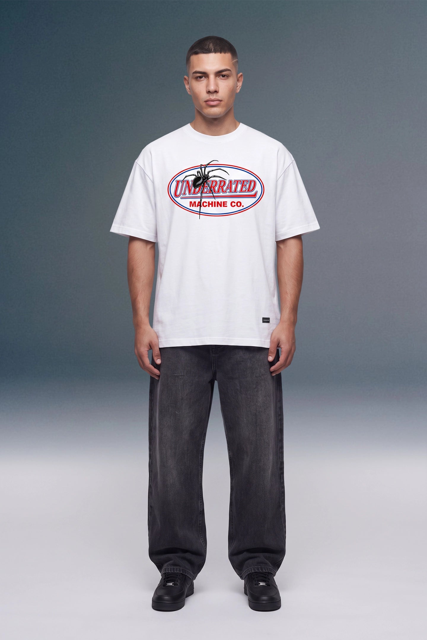 A male model stands against a gray studio background wearing an oversized white graphic T-shirt with a red and black 'Unbranded Machine Co.' logo, dark gray baggy jeans, and black shoes.