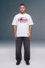 A male model stands against a gray studio background wearing an oversized white graphic T-shirt with a red and black 'Unbranded Machine Co.' logo, dark gray baggy jeans, and black shoes.