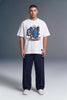 A man modeling a white short-sleeve graphic t-shirt with a blue and black motorcycle print, paired with dark blue jeans and white sneakers, set against a gray studio background.
