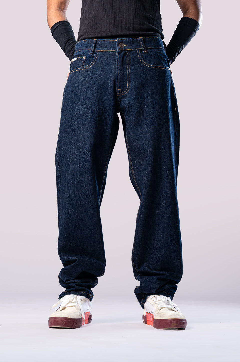 Full-body front view of a person wearing dark-wash blue baggy jeans with a black ribbed top and black arm sleeves, with their hands in their pockets.