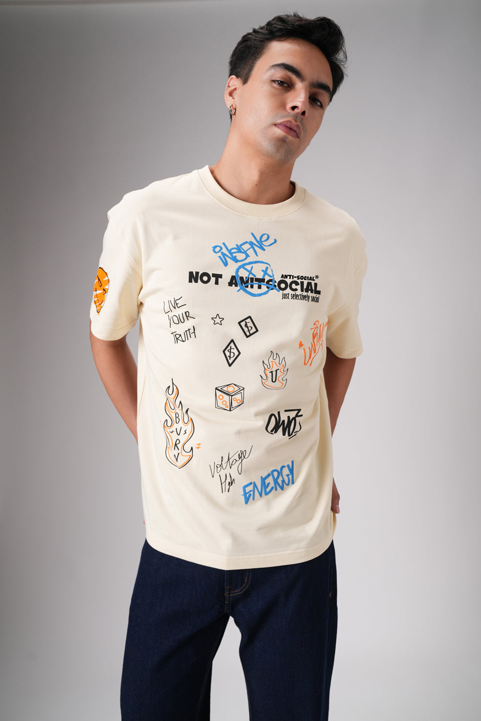 Man looking to the side wearing beige all‑over printed graphic t‑shirt and dark jeans in studio shot.
