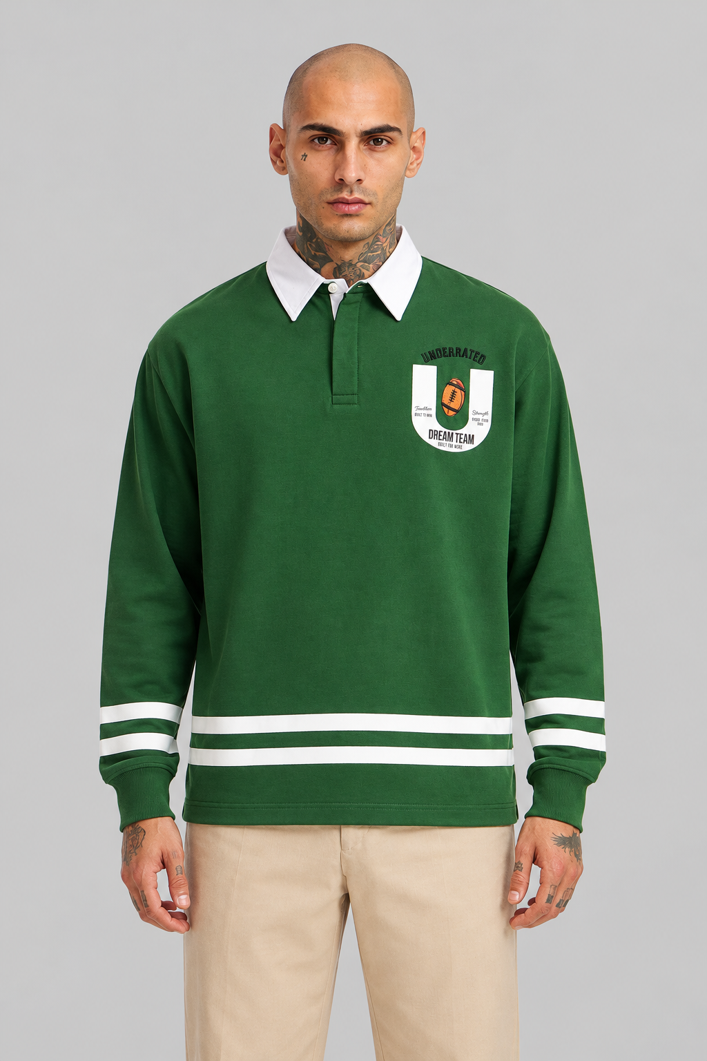 A man wearing a green long-sleeve rugby-style pullover with a white collar, white chest patch, and white stripes on the sleeves, paired with beige pants, against a solid gray studio background.