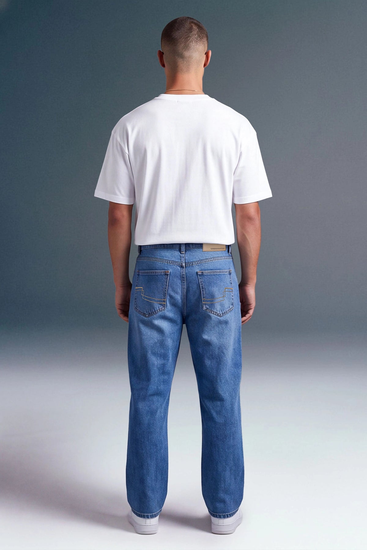 A man viewed from behind wearing a white short-sleeve t-shirt and blue jeans, standing against a dark gray studio background.