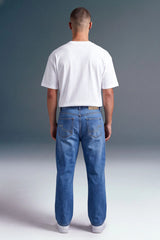 A man viewed from behind wearing a white short-sleeve t-shirt and blue jeans, standing against a dark gray studio background.