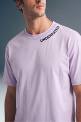 Medium close-up shot from the chest up of a man looking away, wearing a light purple T-shirt featuring subtle black collar text along the left side of the neckline.