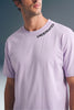 Medium close-up shot from the chest up of a man looking away, wearing a light purple T-shirt featuring subtle black collar text along the left side of the neckline.
