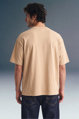 Rear view from the waist up of a man looking to the side, showcasing the back fit and shoulder structure of a loose beige T-shirt paired with dark blue jeans.