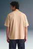 Rear view from the waist up of a man looking to the side, showcasing the back fit and shoulder structure of a loose beige T-shirt paired with dark blue jeans.