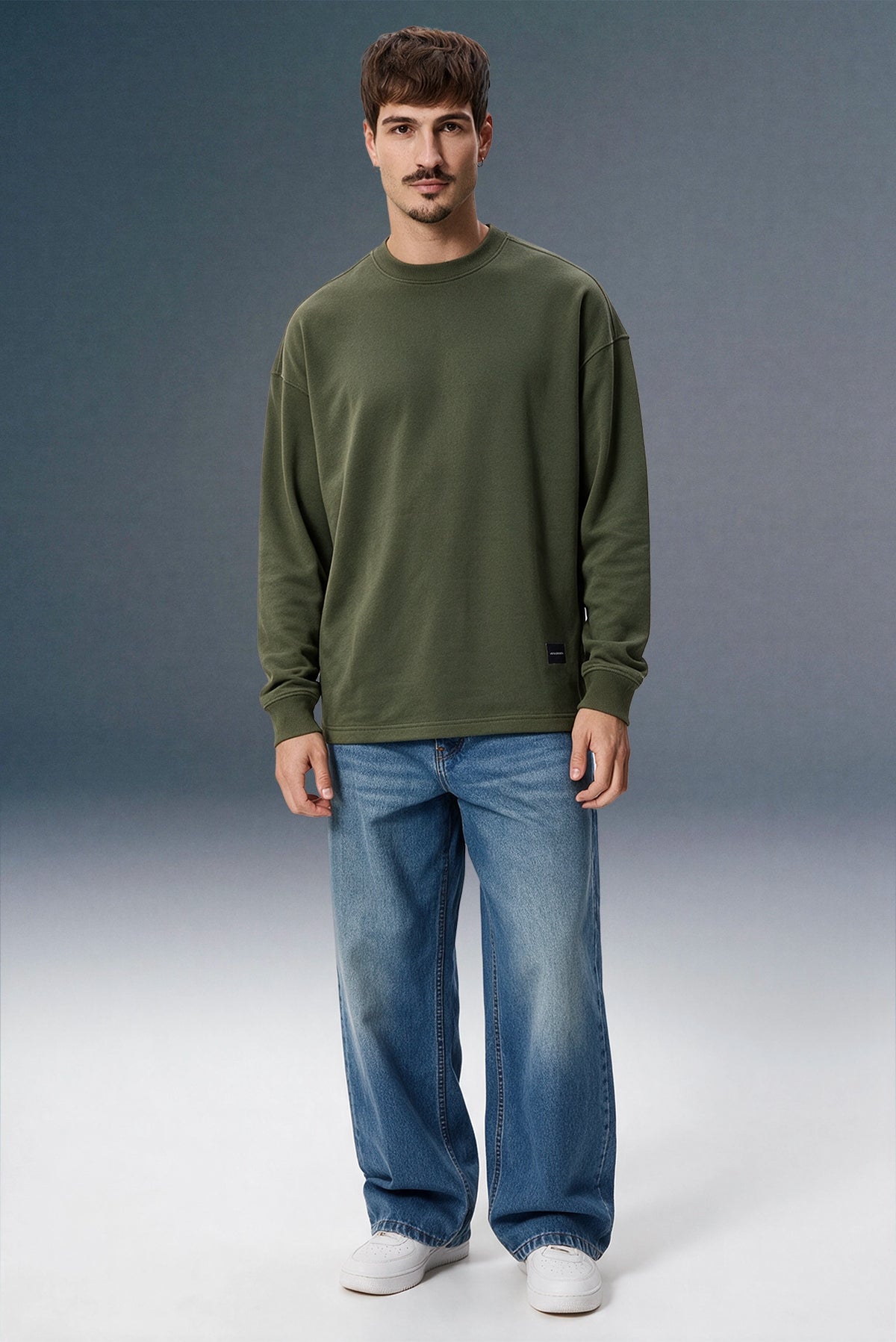 A man wearing an olive green long-sleeve crewneck sweatshirt and blue jeans, standing against a gray studio background.