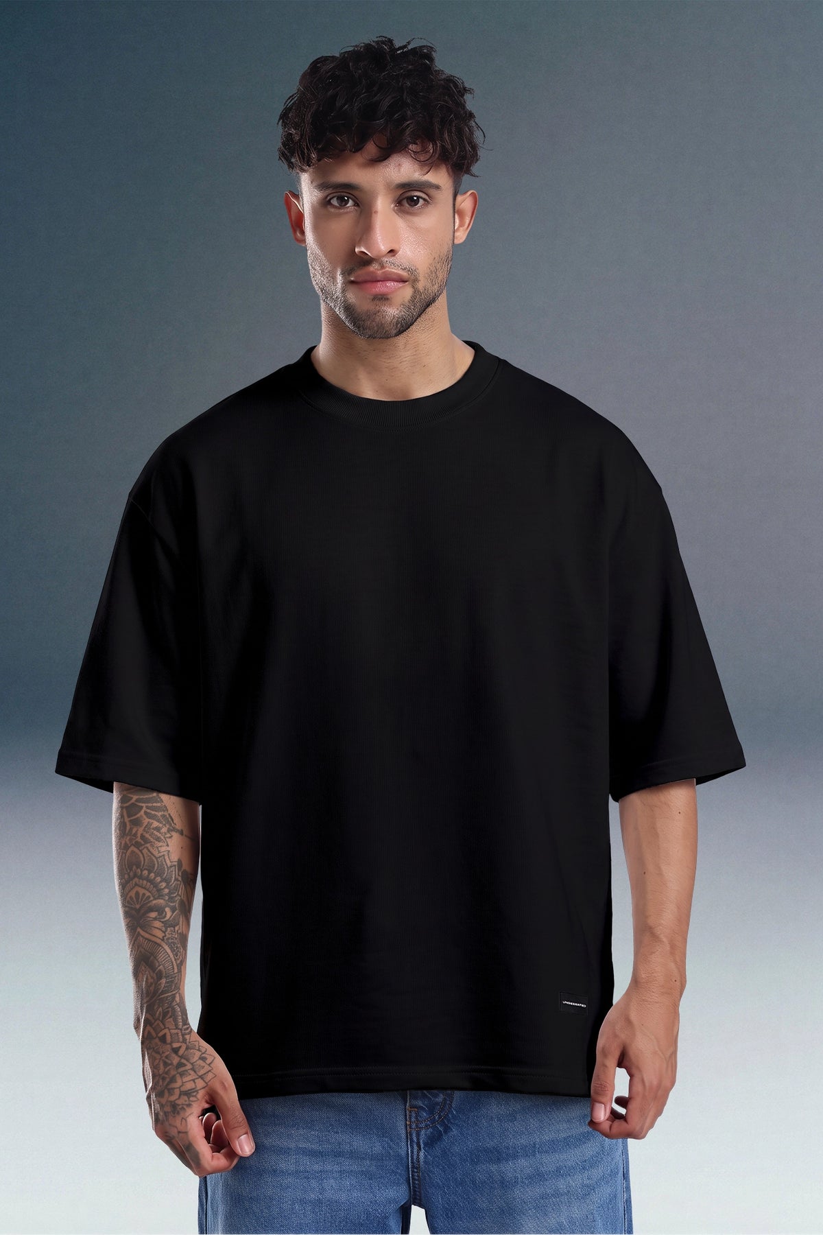 A man wearing an oversized black short-sleeve t-shirt and blue jeans, standing against a gray studio background.