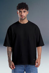 A man wearing an oversized black short-sleeve t-shirt and blue jeans, standing against a gray studio background.