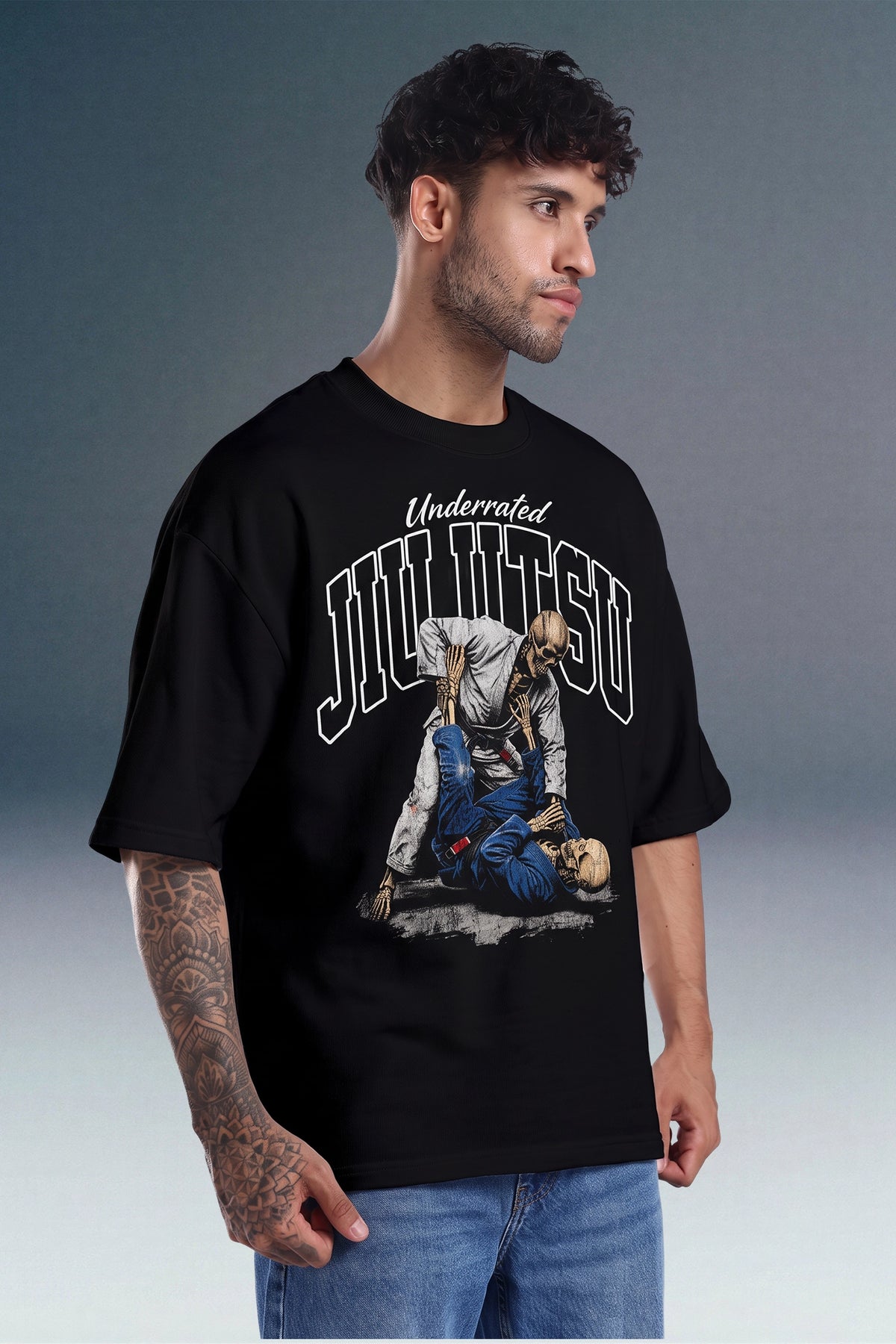 A man wearing a black graphic t-shirt with a white and blue 'Underdogged Jiu Jitsu' print, paired with blue jeans, set against a gray studio background.
