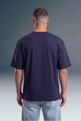 A man viewed from behind wearing a dark navy blue short-sleeve t-shirt and light blue jeans, standing against a dark gray gradient studio background.