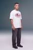 A man modeling a white short-sleeve t-shirt with a red and black 'ULTRAFOX DAKARUS CITY' graphic on the chest, paired with dark gray jeans and black shoes, against a studio backdrop.