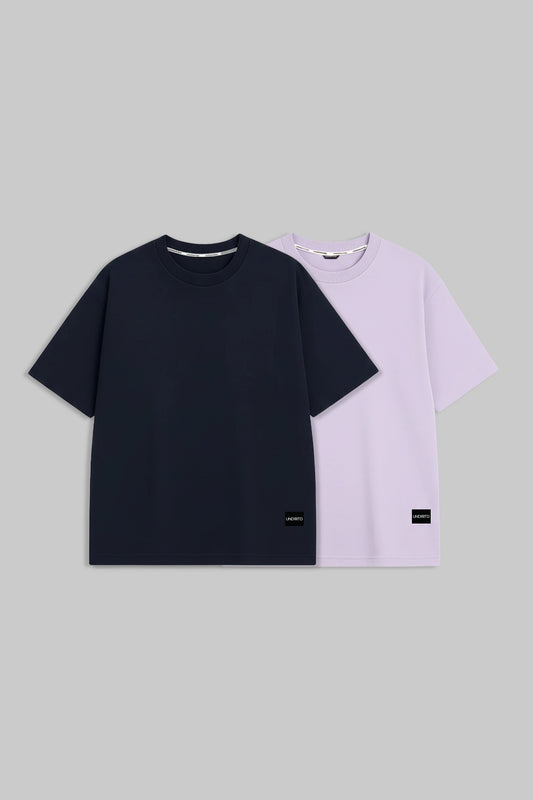 Two short-sleeve crewneck T-shirts displayed flat against a solid gray background. The front shirt is solid black, and the shirt behind it is a light lavender purple, both featuring small black rectangular labels near the bottom hem.