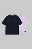 Two short-sleeve crewneck T-shirts displayed flat against a solid gray background. The front shirt is solid black, and the shirt behind it is a light lavender purple, both featuring small black rectangular labels near the bottom hem.