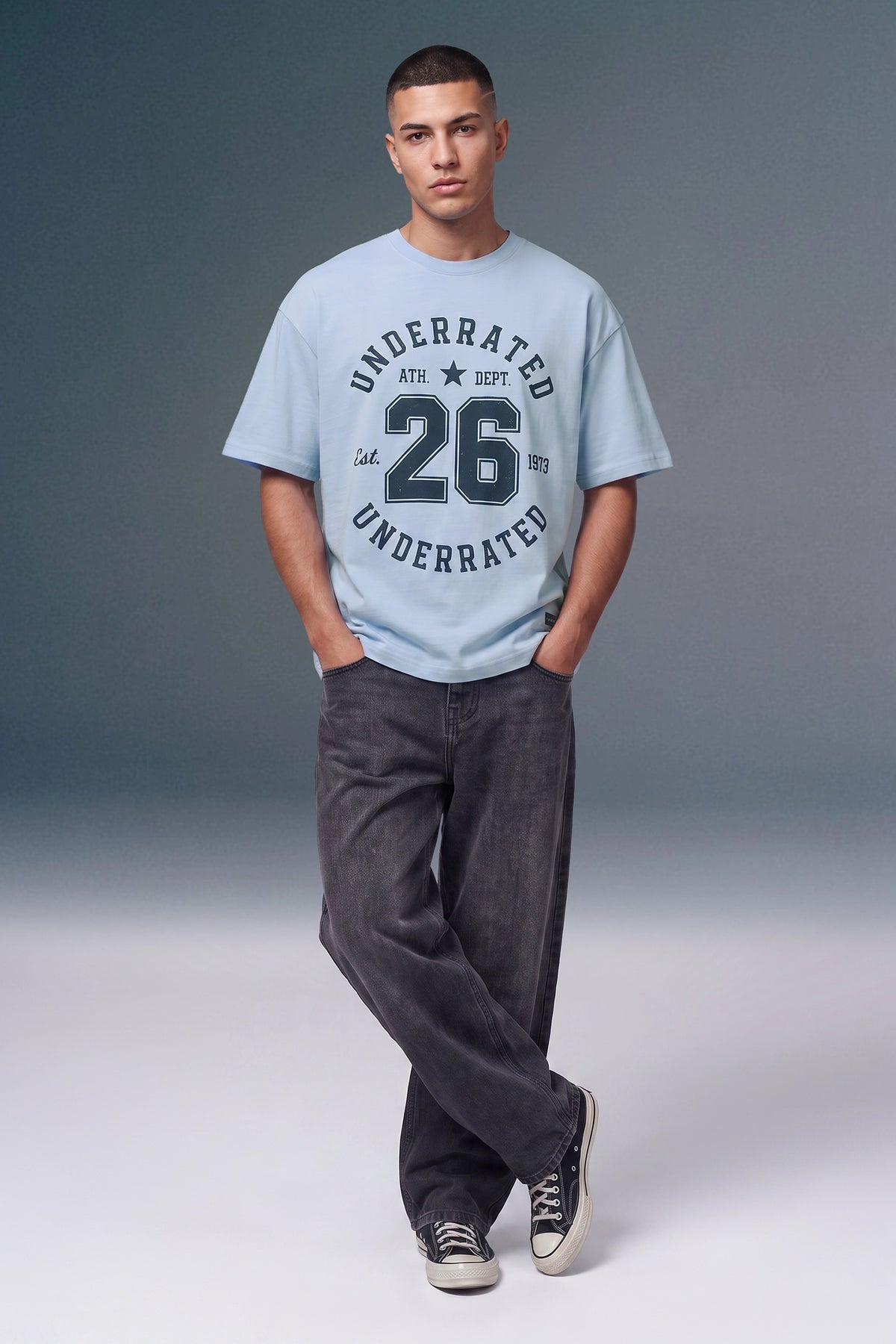 A man modeling a light blue graphic t-shirt with 'UNDERRATED 26 EST.' printed on the front, paired with dark gray jeans and black sneakers, against a gray studio background.