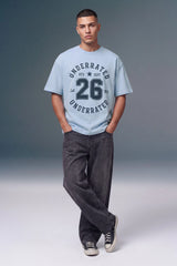 A man modeling a light blue graphic t-shirt with 'UNDERRATED 26 EST.' printed on the front, paired with dark gray jeans and black sneakers, against a gray studio background.