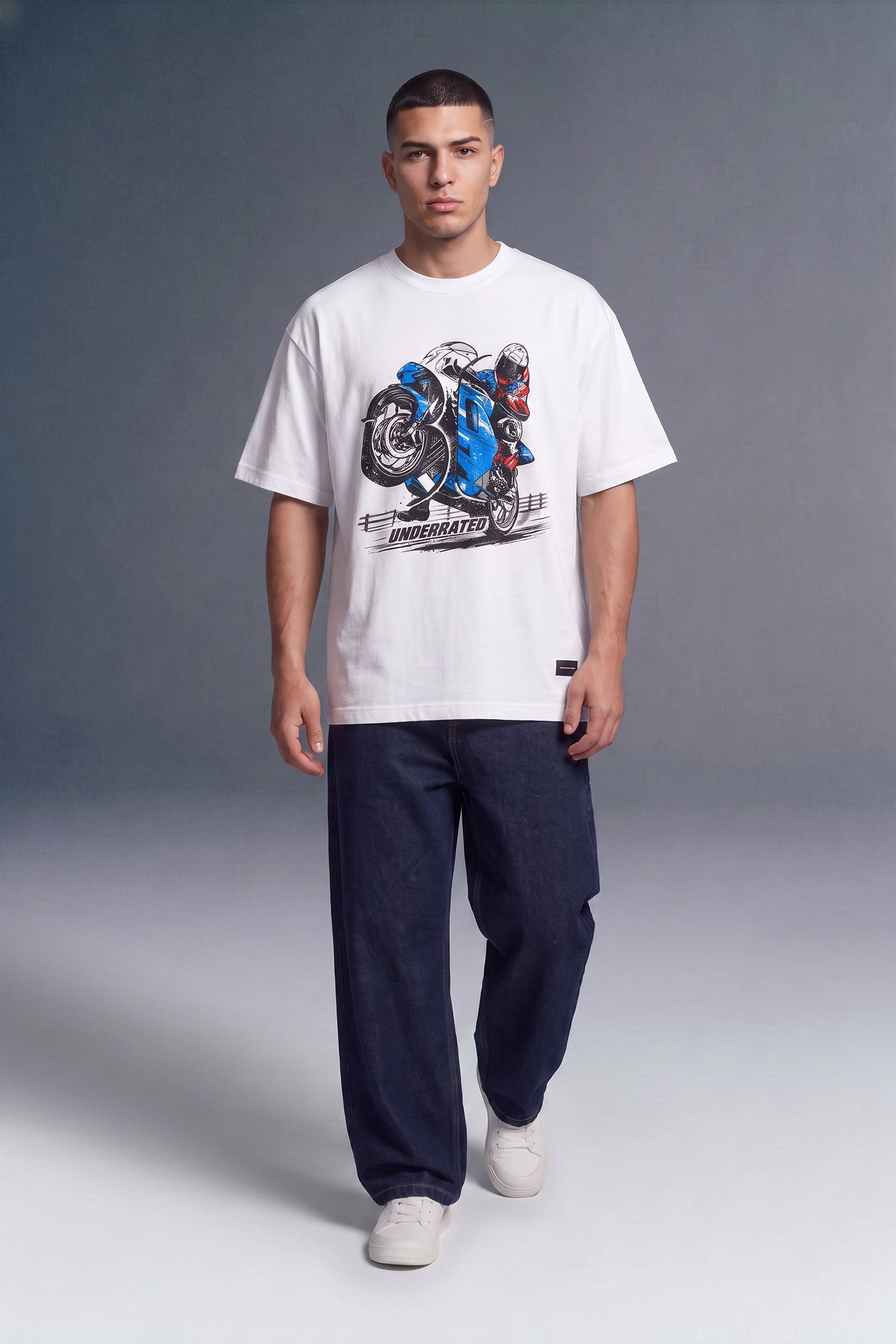 A man modeling a white short-sleeve graphic t-shirt with a blue and black motorcycle print, paired with dark navy pants and white sneakers, against a gray studio background.
