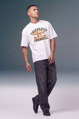 A man modeling a white graphic t-shirt with a distressed orange and green print that reads 'UNDERDATED BUT STYLIZED UNDERDATED', paired with dark gray jeans and black shoes, against a studio background.