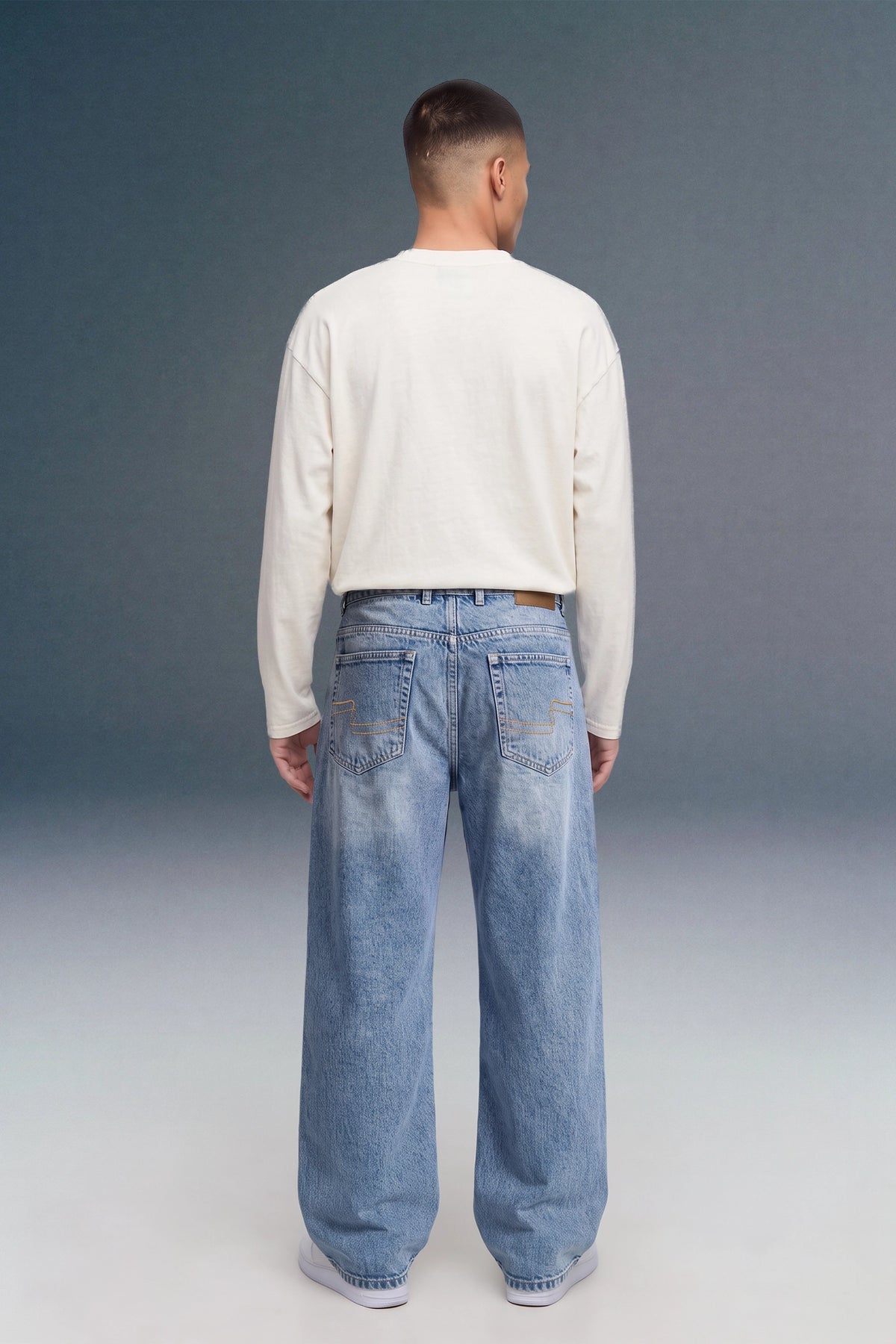A man viewed from behind wearing a cream long-sleeve shirt and light-wash blue wide-leg jeans against a dark gray studio background.